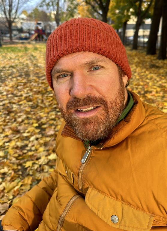 Luke Anderson is posing for a selfie." longdesc="Luke is wearing a red beanie and a beige jacket. He is sitting in a park with leaves covering the ground and trees in the background.